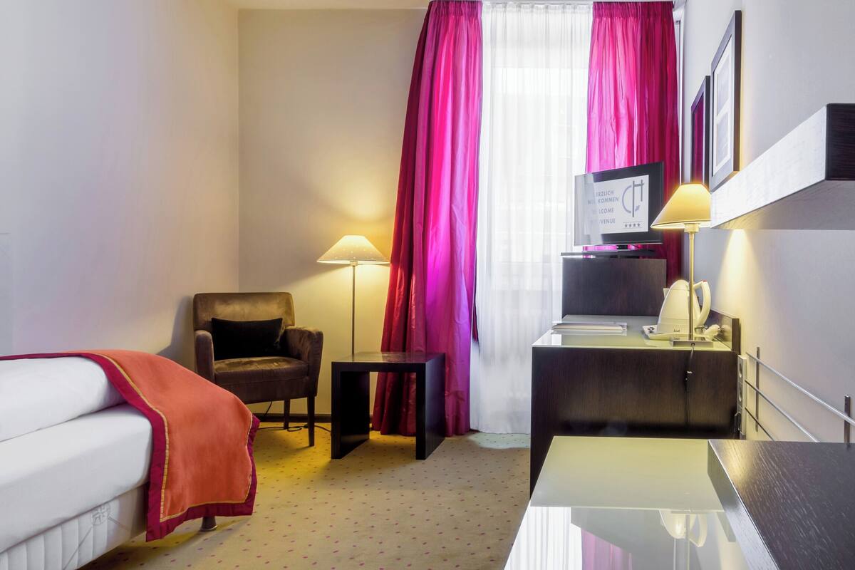 Habitación de hotel con cama, escritorio, silla, televisor, lámpara y ventana con cortinas.