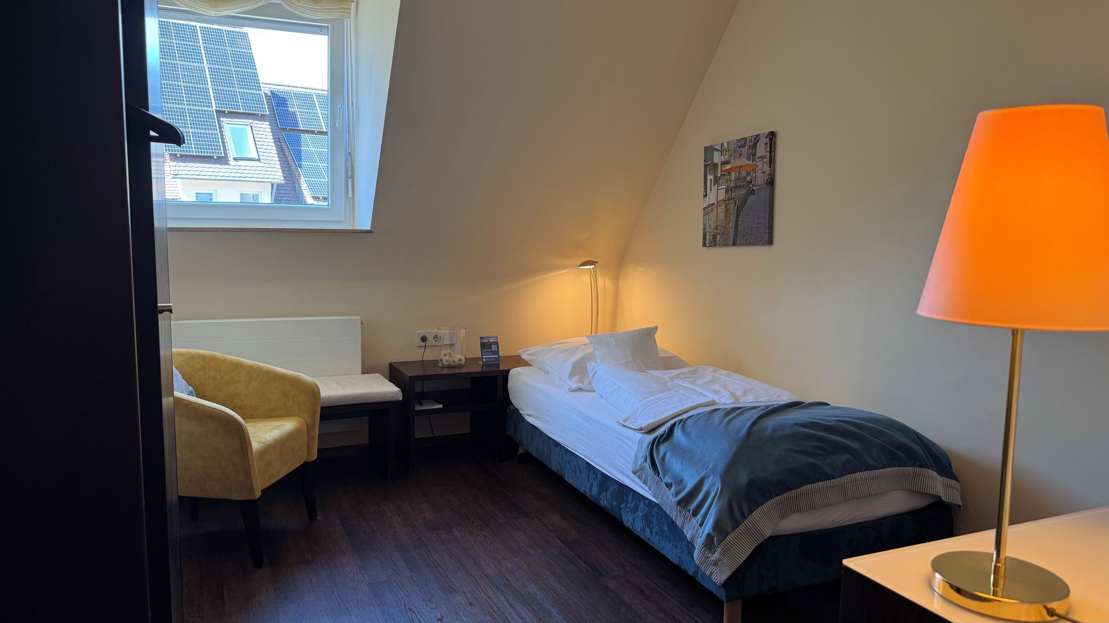 A bedroom with a bed, bedside table, lamp, chair, and a window with a view of buildings.