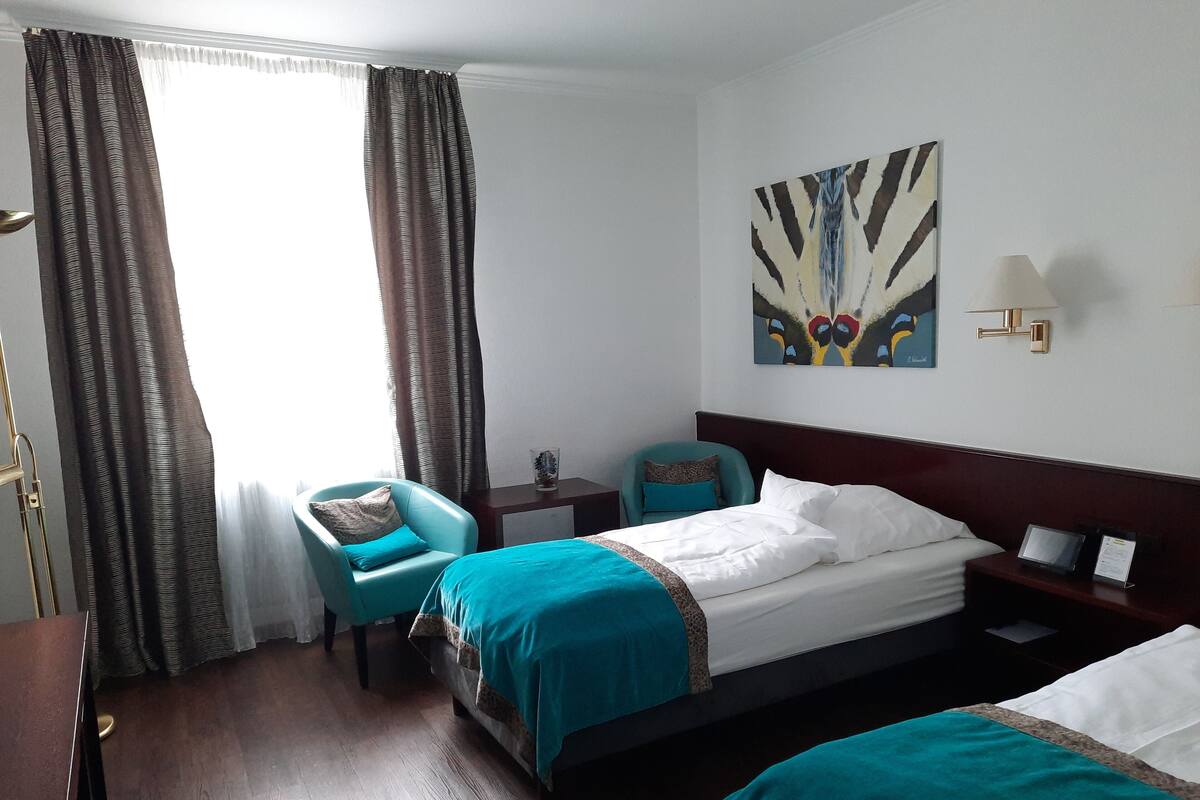 Habitación de hotel con dos camas, un escritorio, una silla, un ventanal con cortinas y una lámpara de pared.