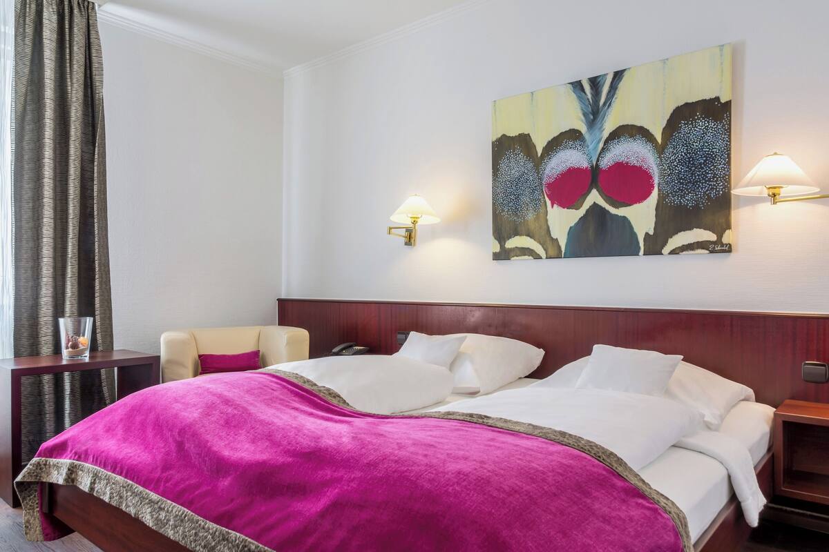 Una habitación de hotel con una cama, una silla, una mesita de noche, una lámpara y un cuadro en la pared.