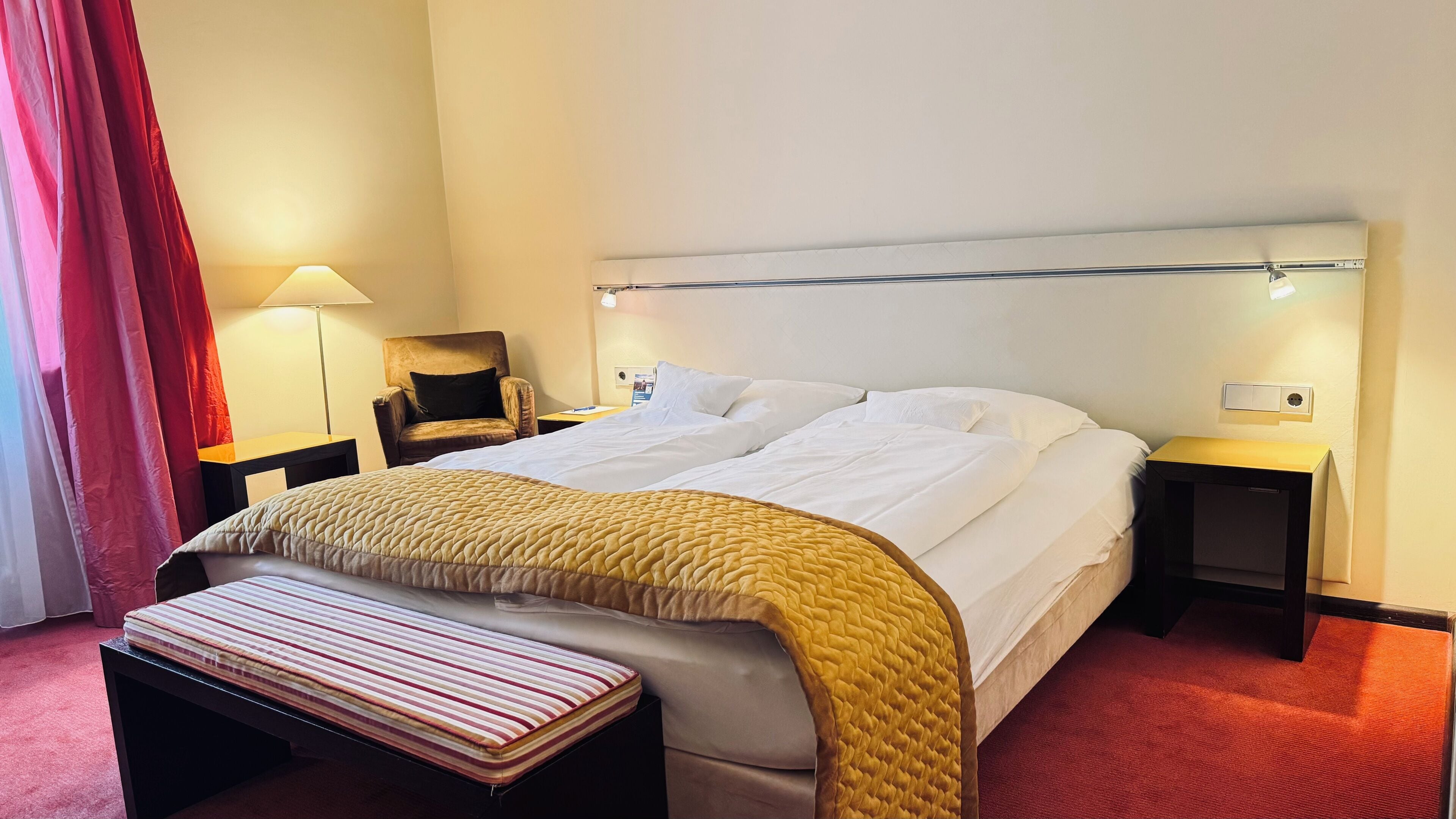 Quarto de hotel com cama, mesas de cabeceira, uma cadeira, um banco e uma luminária.