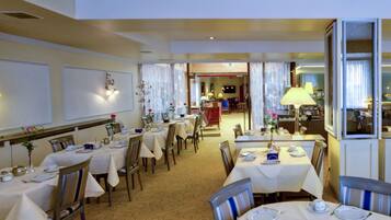 A restaurant with multiple tables and chairs, a chandelier, and a view into the kitchen area.
