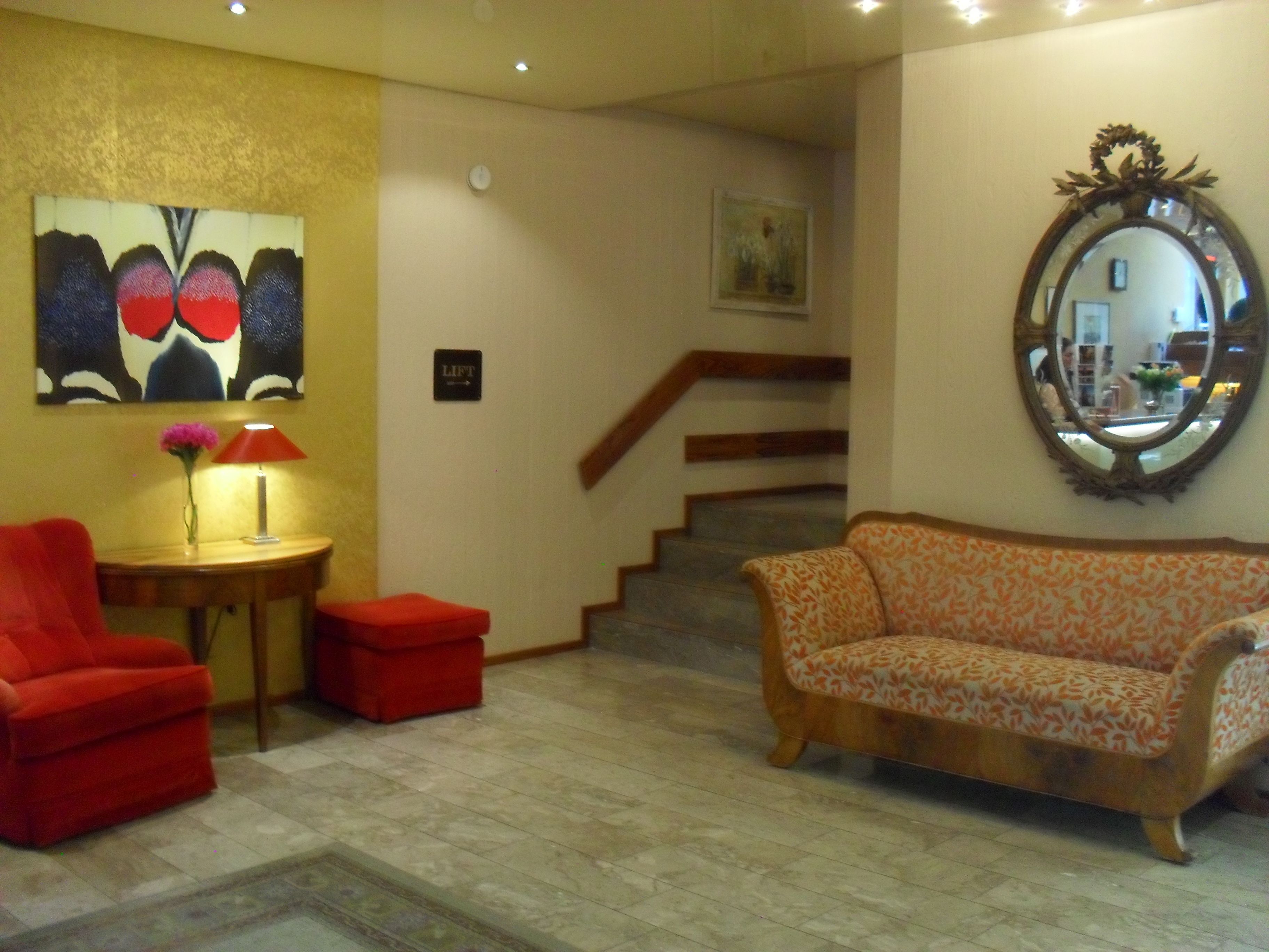 Eine Lobby mit einem roten Sessel, einer gemusterten Couch, einem kleinen Tisch mit Lampe, einem Gemälde, einem Spiegel und einer Treppe.