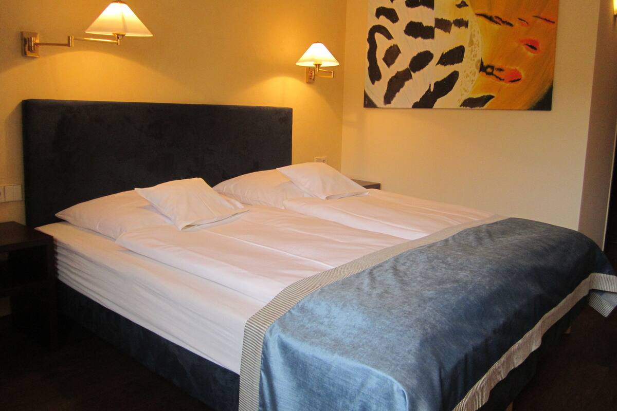 Una cama bien hecha con sábanas blancas y un detalle azul, dos lámparas de noche y un cuadro en la pared.