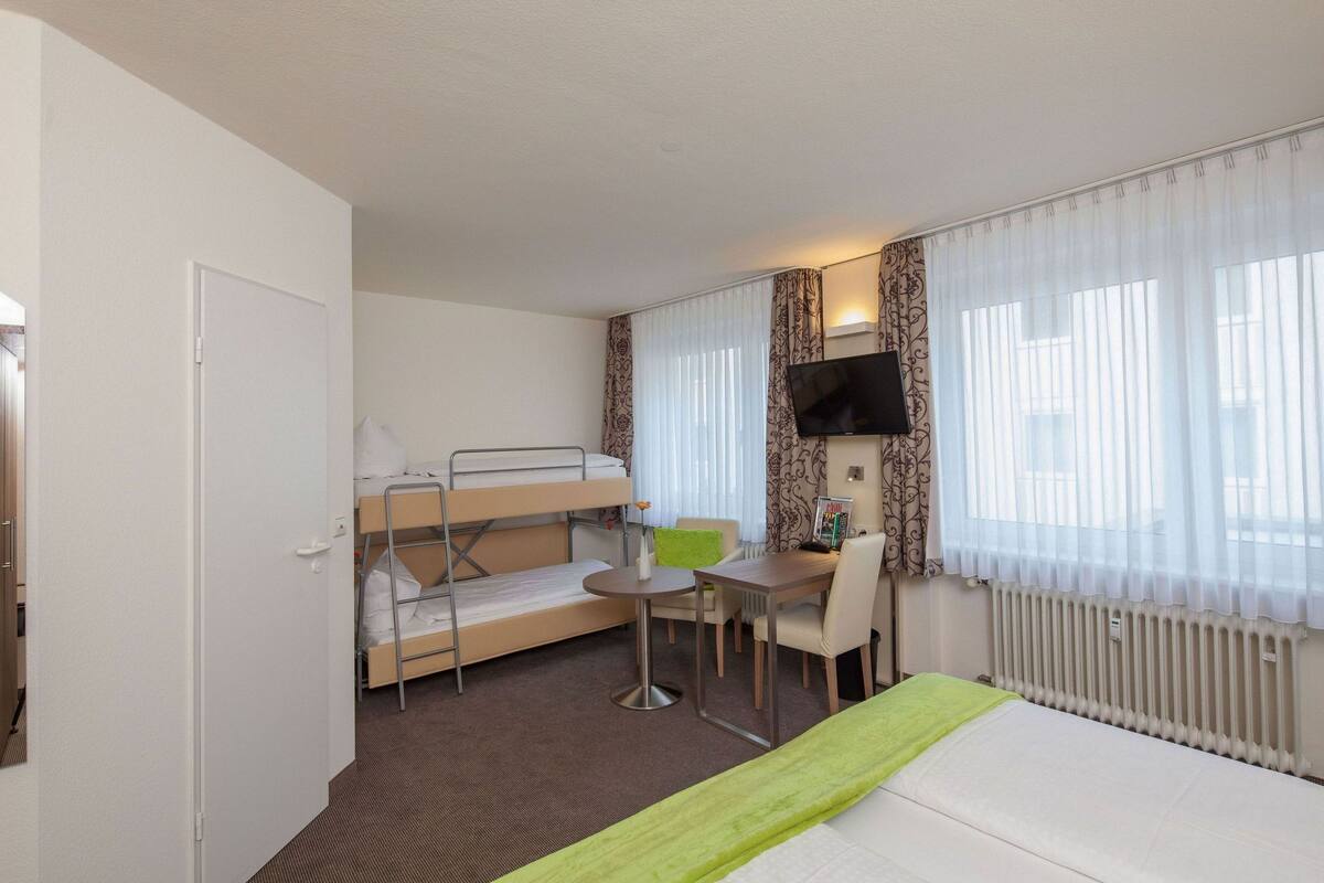 Une chambre d’hôtel avec des lits superposés, une petite table avec une chaise, un téléviseur et une fenêtre avec des rideaux légers.