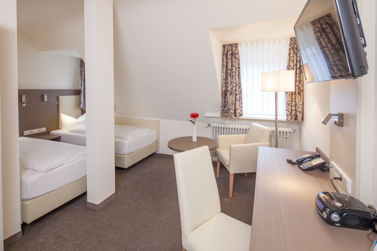 Une chambre d’hôtel avec deux lits, un bureau avec un téléphone et une radio, une chaise, une petite table et un radiateur.