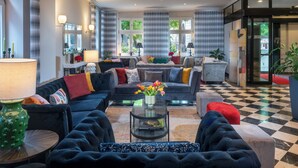 A spacious living room with a checkered floor, a sofa set, a coffee table with a vase of flowers, and a view of the outdoors through large windows. - Sorat Hotel Cottbus (Cottbus)