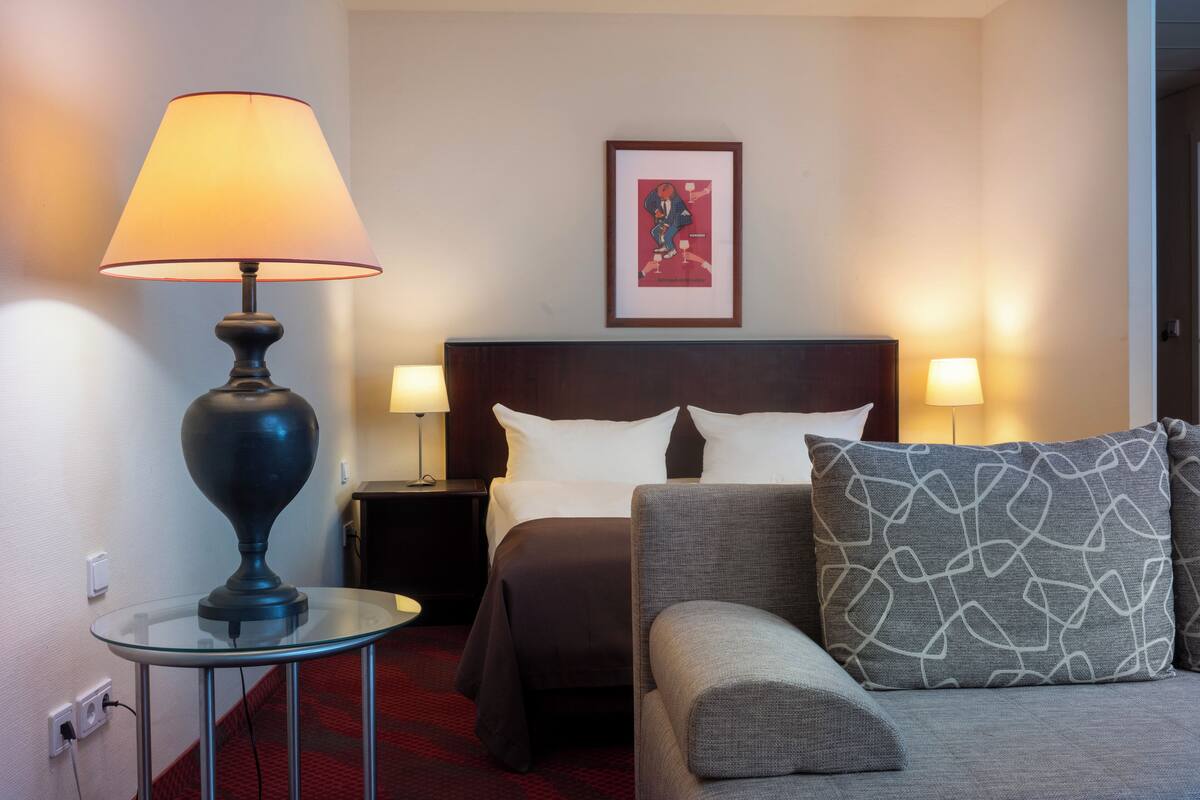A hotel room with a bed, two bedside lamps, a glass table with a lamp, a sofa, and a framed picture on the wall.