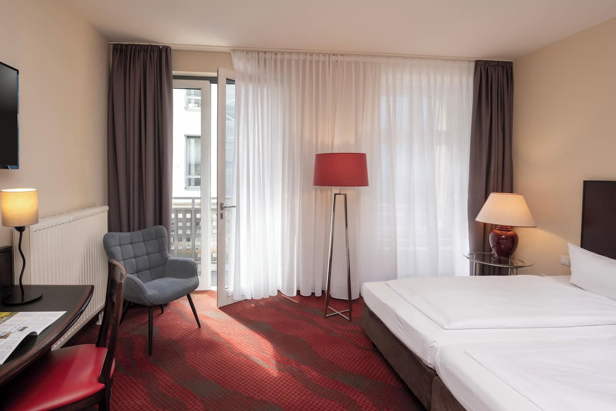 A hotel room with a bed, a desk with a chair, a standing lamp, and a view of the outside through a window with curtains.