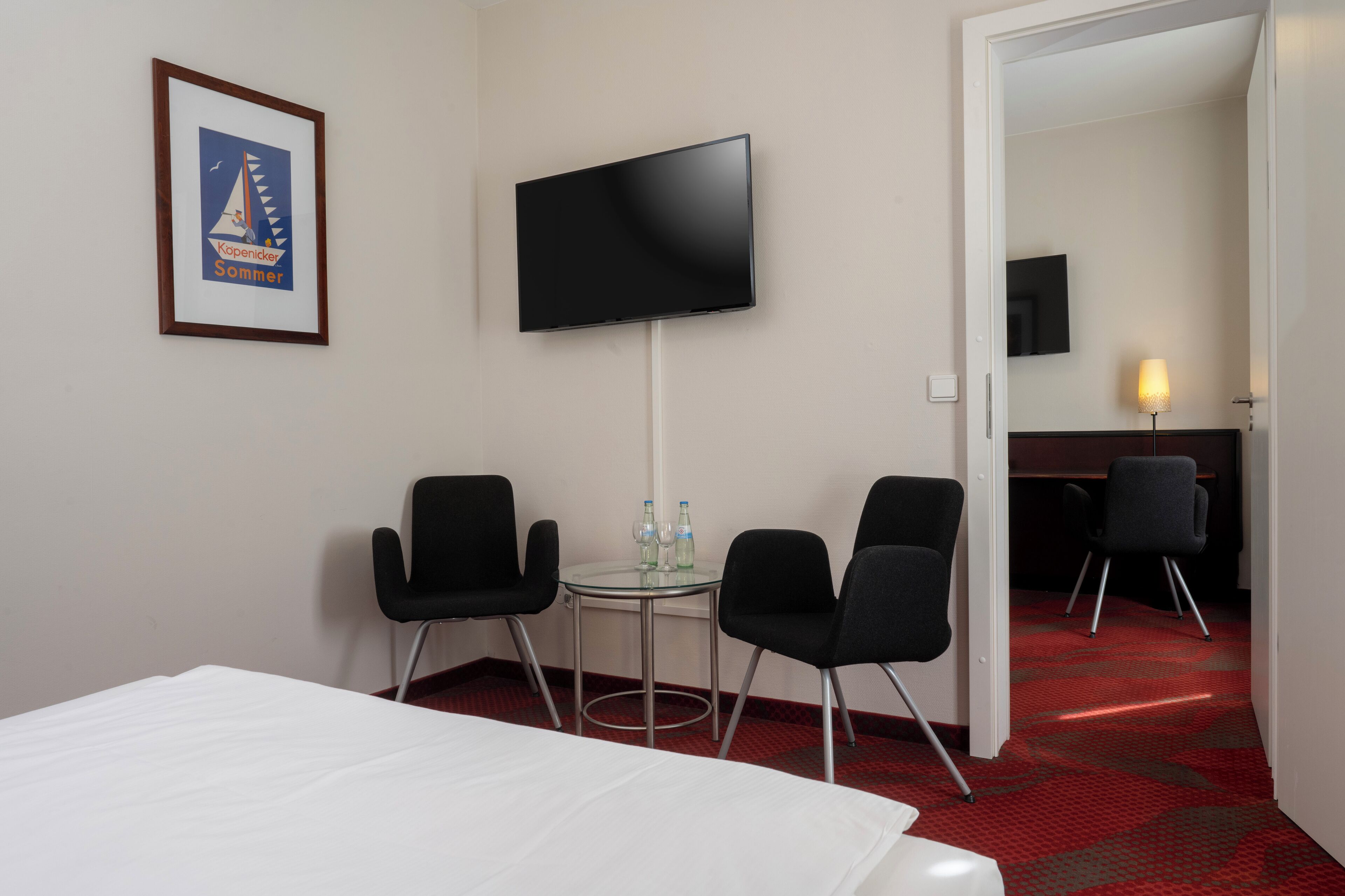 Habitación de hotel con una cama, dos sillas negras, una mesita con botellas de agua, un televisor montado en la pared y un cuadro enmarcado.