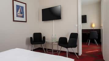 Habitación de hotel con una cama, dos sillas negras, una mesita con botellas de agua, un televisor montado en la pared y un cuadro enmarcado.