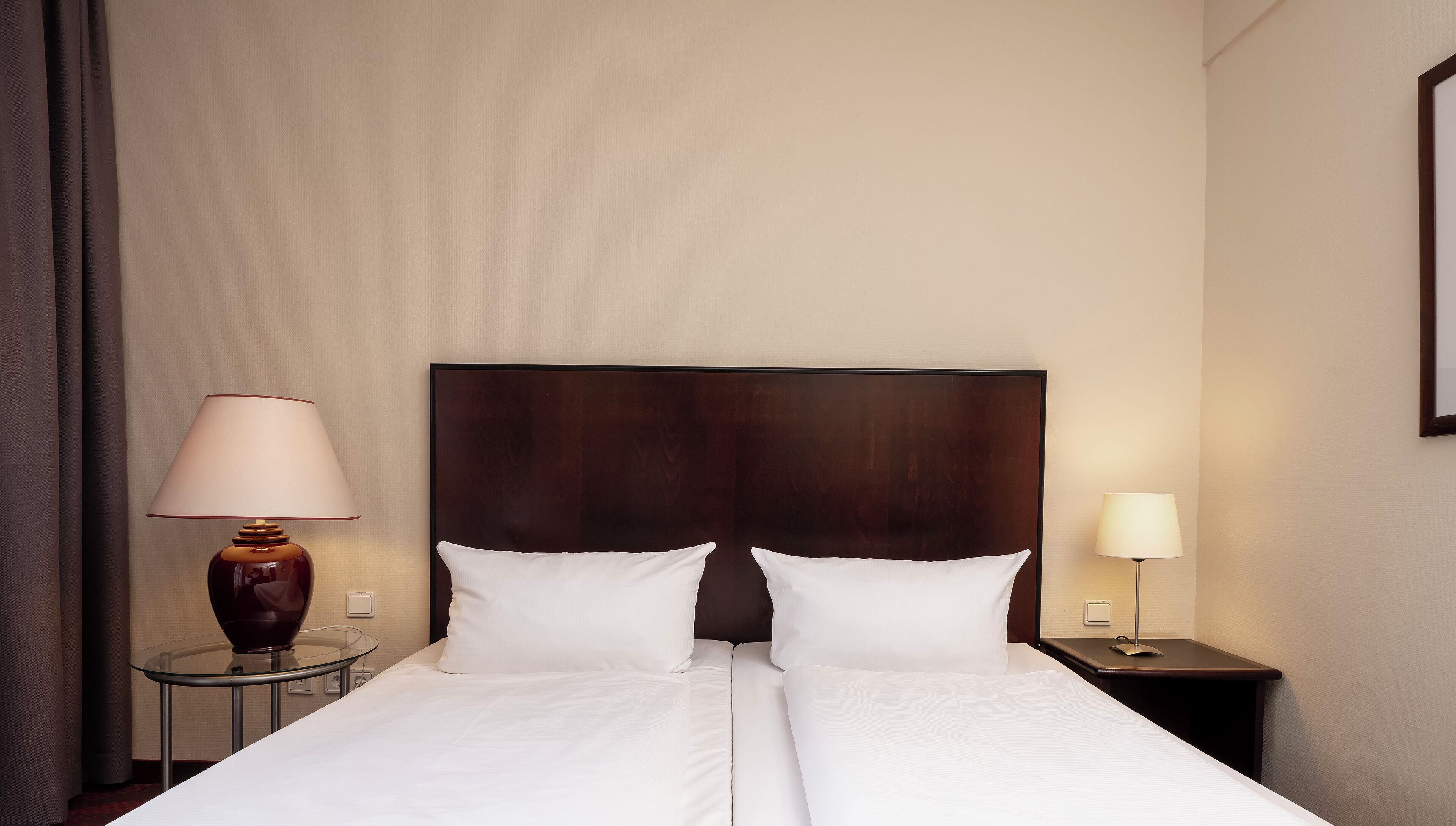 A double bed with white linens, a dark wood headboard, two bedside tables with lamps, and a plain wall.