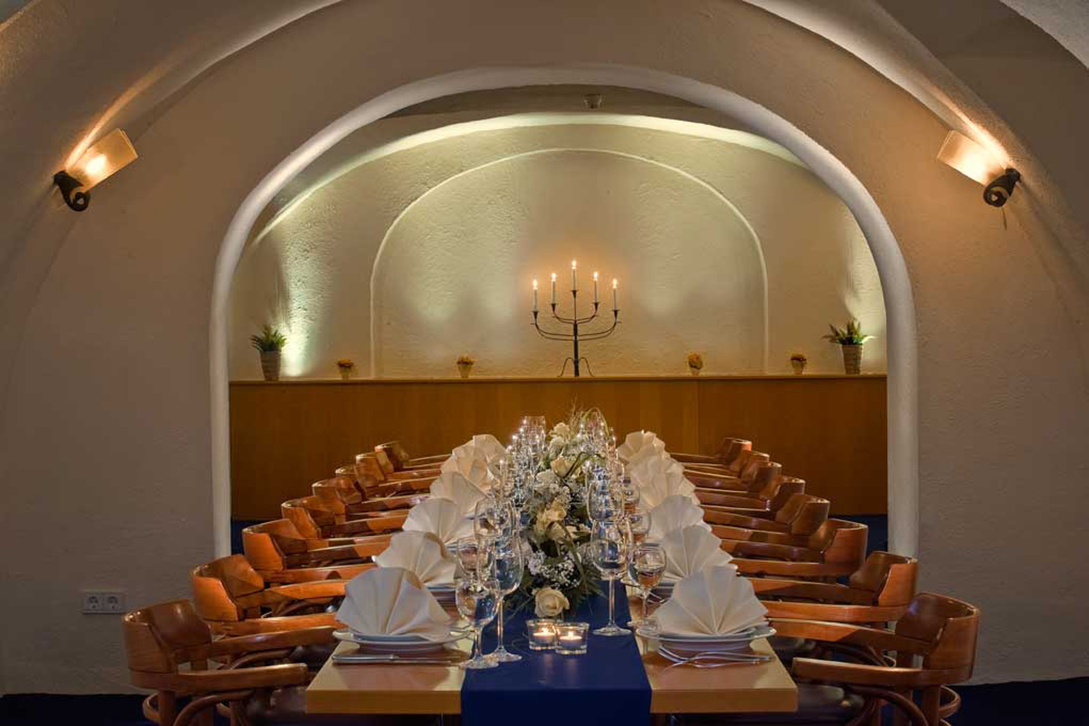 A formal dining room with a long table set for a meal, flanked by wooden chairs, under a semi-circular arched ceiling with wall-mounted lighting.