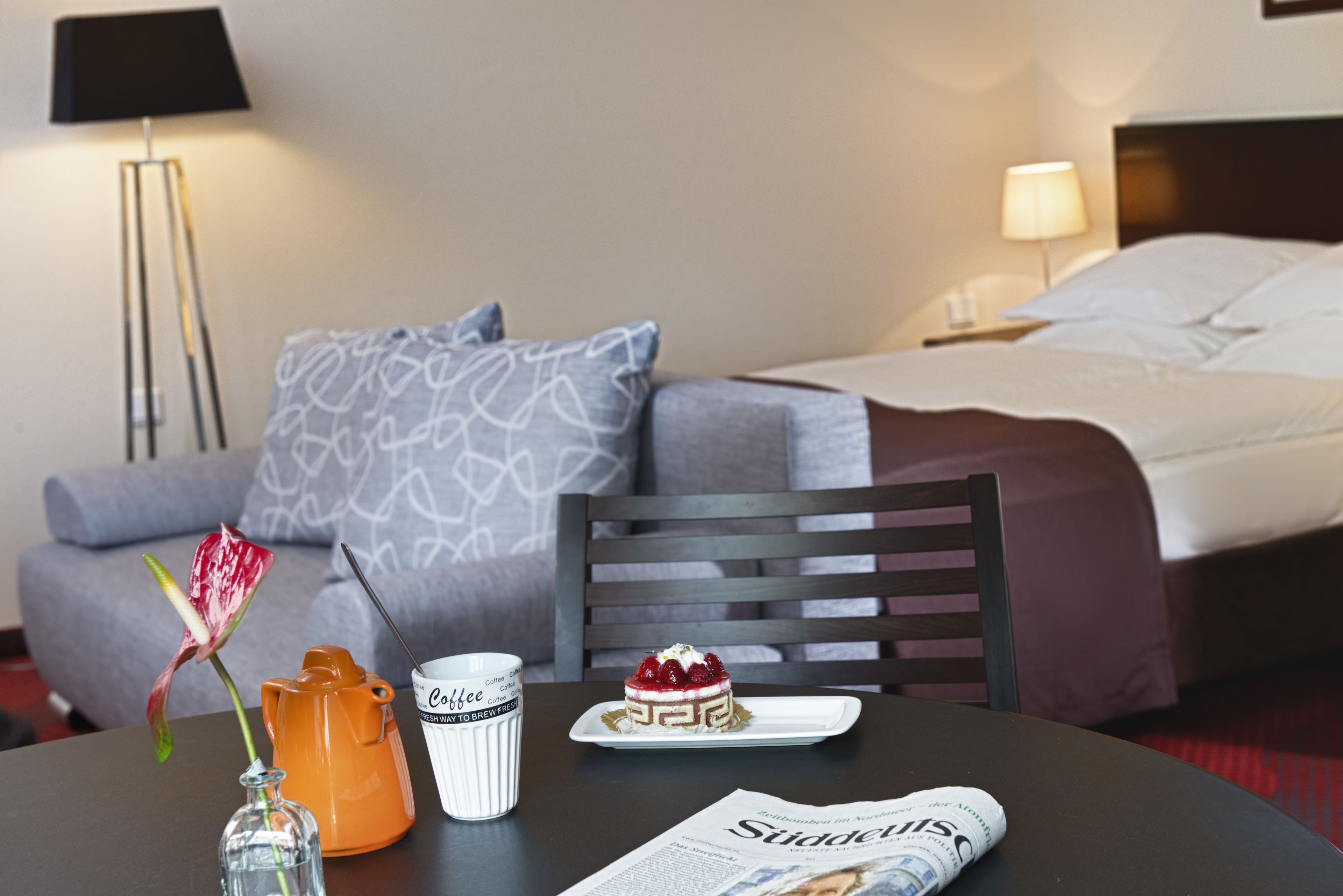 A hotel room with a bed, a sofa, a table with a newspaper, a coffee pot, a vase with a flower, and a plate with a dessert.
