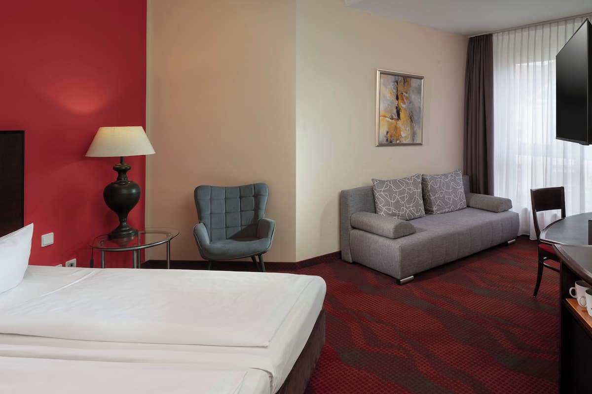 A hotel room with a bed, a sofa, a chair, a TV, and a lamp.