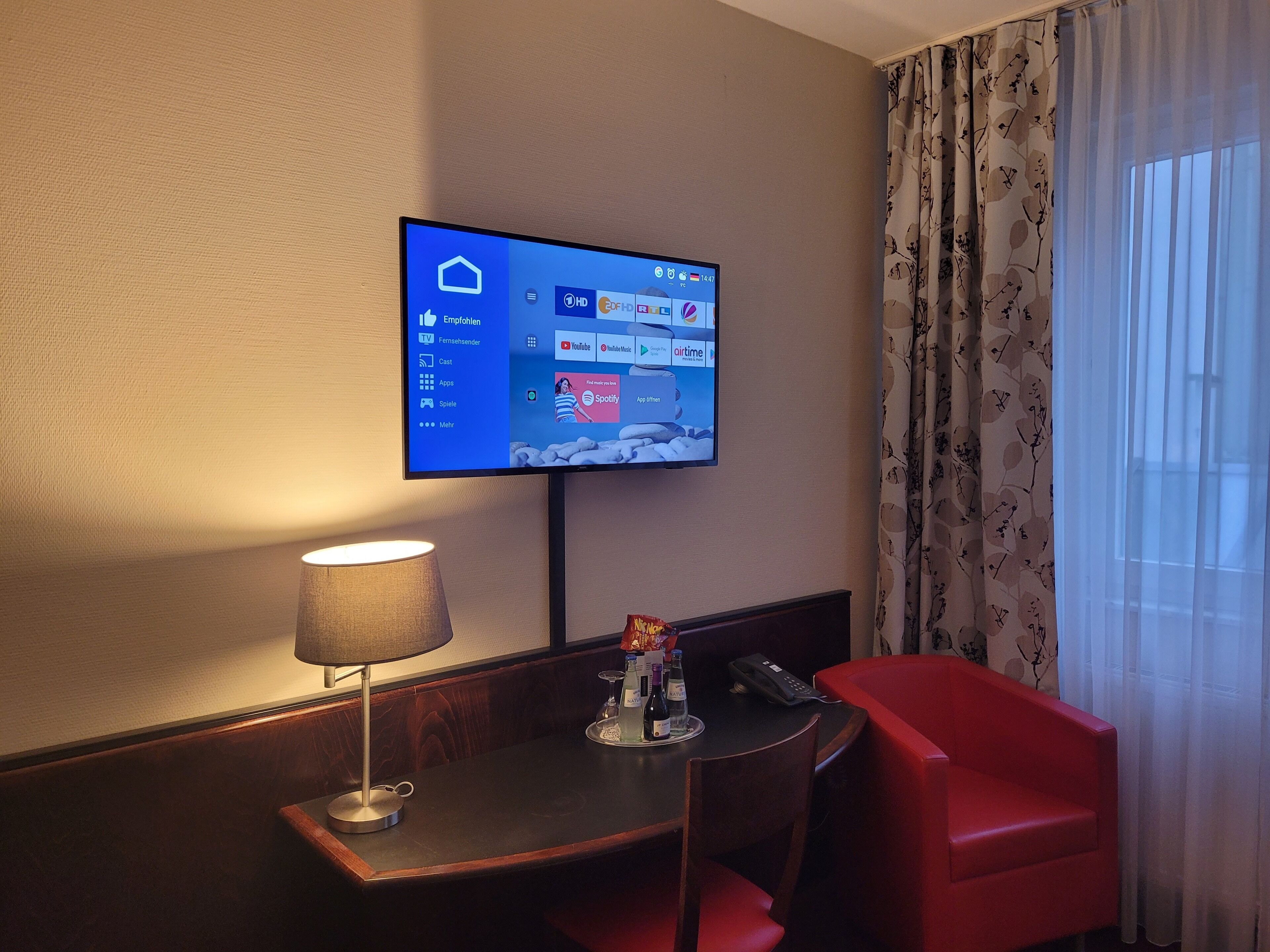 Une chambre d’hôtel équipée d’un téléviseur à écran plat fixé au mur, qui affiche un écran d’accueil avec différentes applications. On y trouve une table de chevet en bois avec une lampe, une chaise rouge et une petite table sur laquelle se trouvent une bouteille et un verre.
