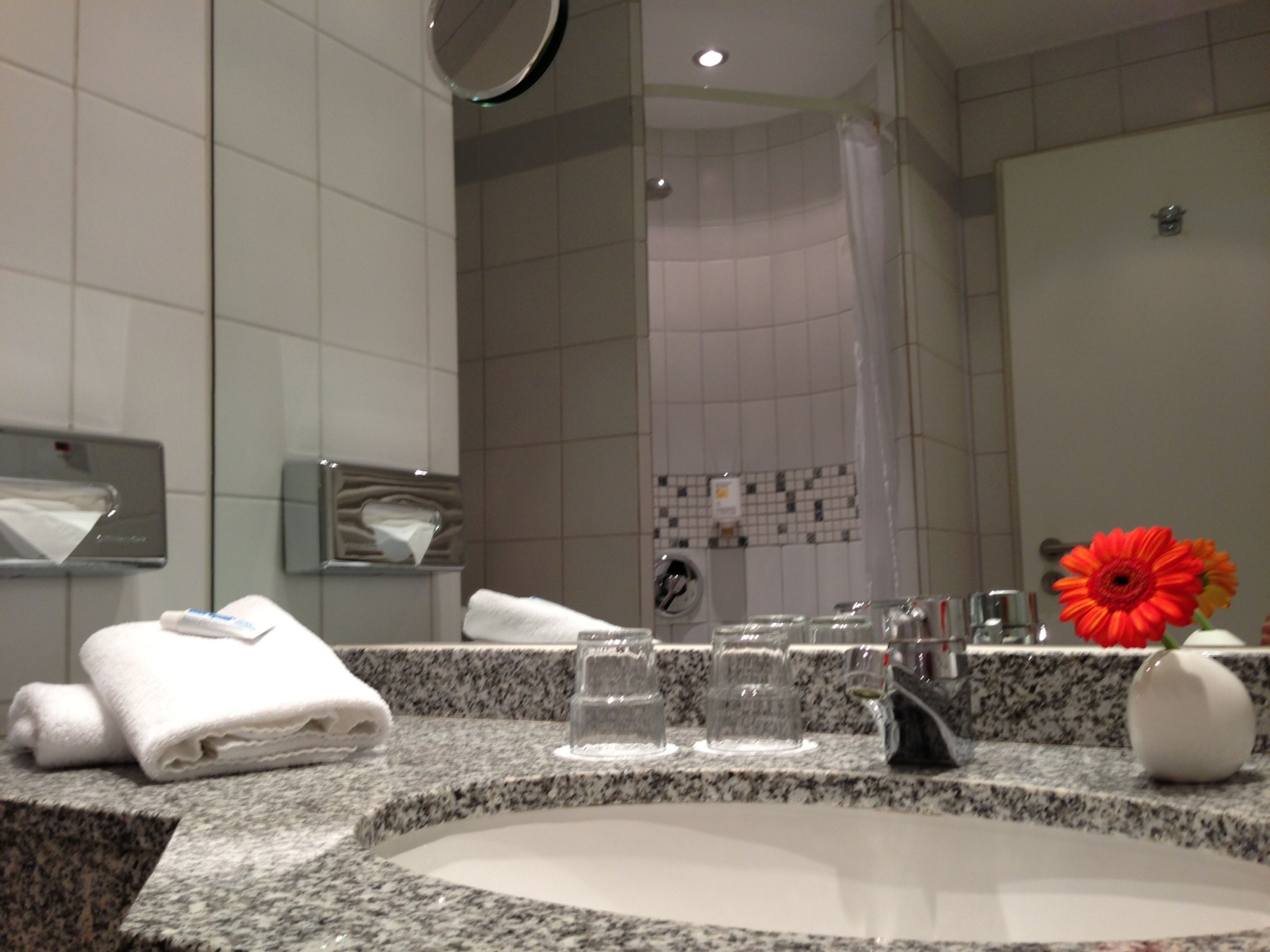 A bathroom with a granite countertop, a sink, a mirror, a towel rack, a glass shower enclosure, and a vase with a red flower.