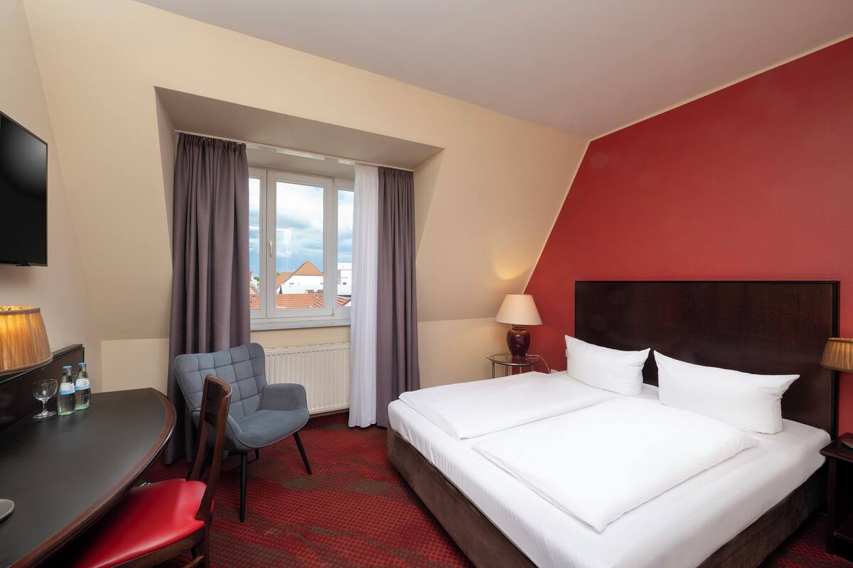 A hotel room with a bed, a desk with a chair, a TV, and a window with curtains.