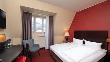 Una habitación de hotel con cama, escritorio con silla, televisión y ventana con cortinas.