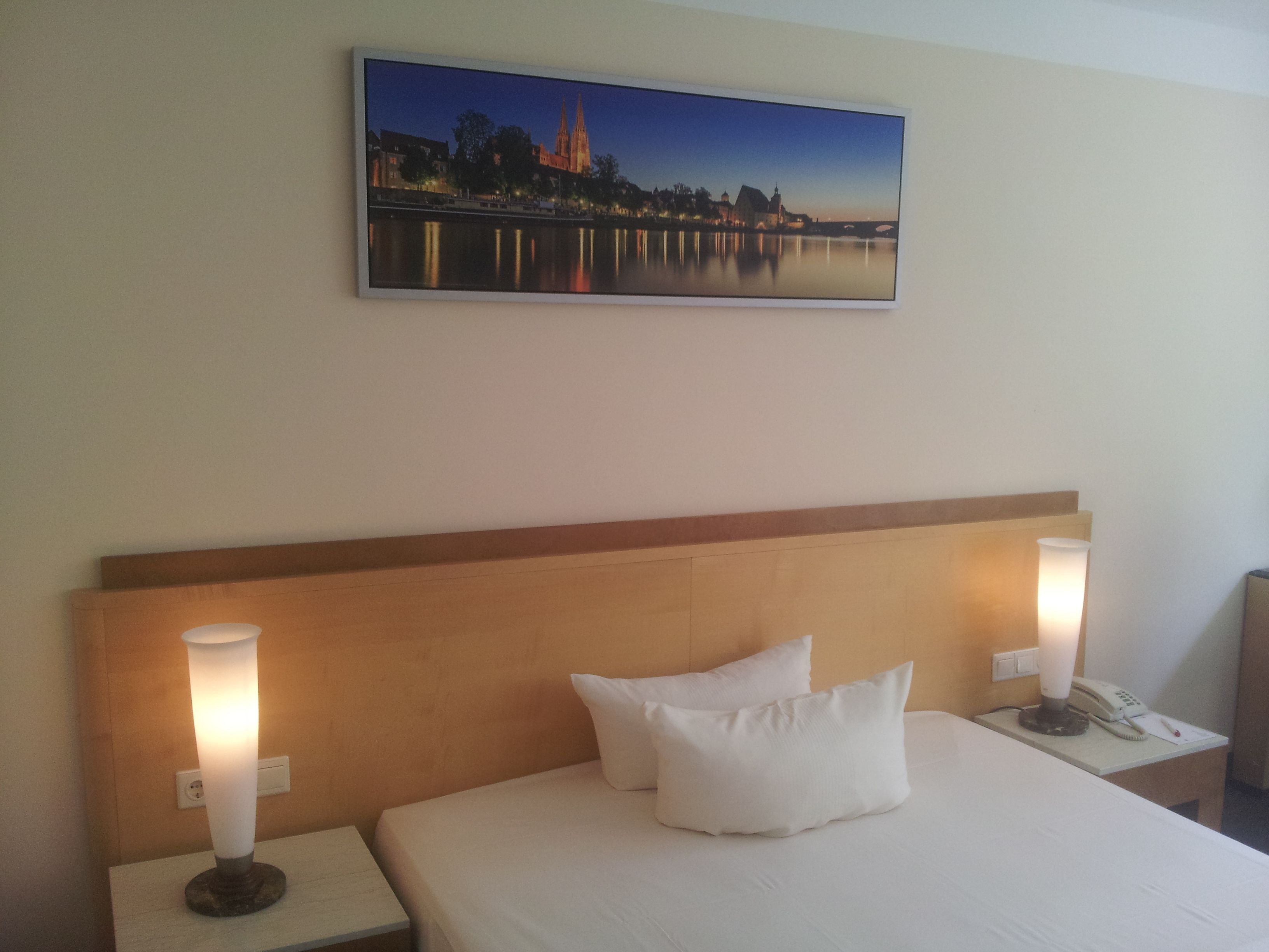 A hotel room with a bed, two bedside lamps, a nightstand, a painting of a cityscape, and a telephone on the nightstand.