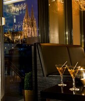 Interior de restaurante com vista para uma catedral pela janela, uma mesa com dois copos de martini e uma pequena planta sobre ela.