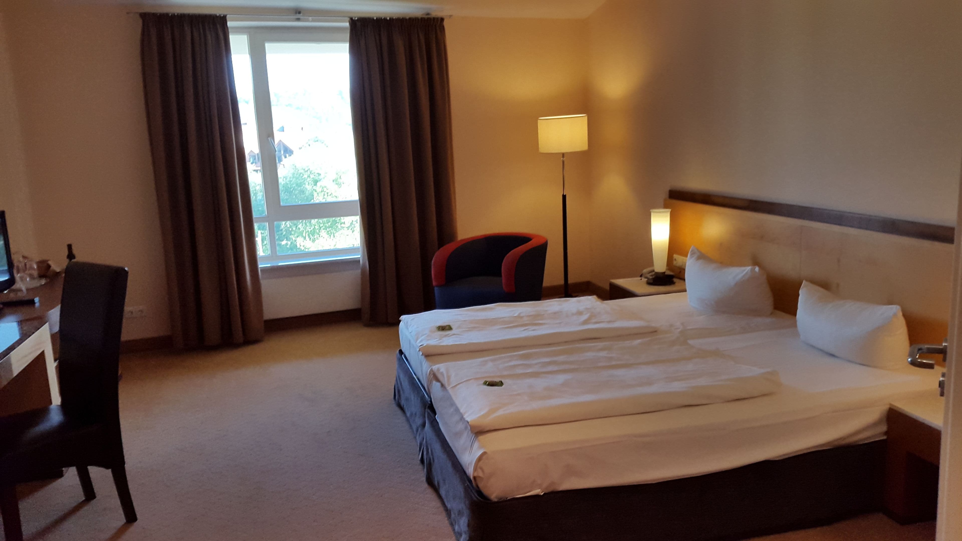 Habitación de hotel con una cama grande, un escritorio, una silla, una lámpara y una ventana con cortinas.