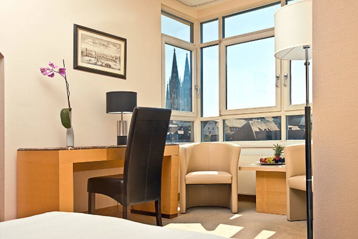 A hotel room with a desk, two chairs, a bed, a lamp, a framed picture, and a window with a view of a building.
