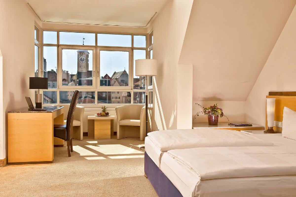 A hotel room with a large window, a desk, a chair, a bed, and a nightstand with a lamp.