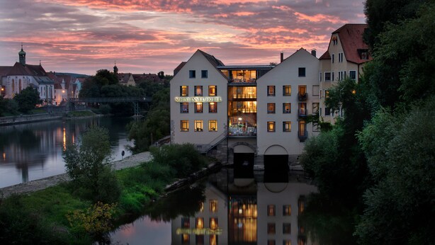 A hotel with a river in front, a bridge, and a sunset sky. - Sorat Insel-Hotel Regensburg (Regensburg)
