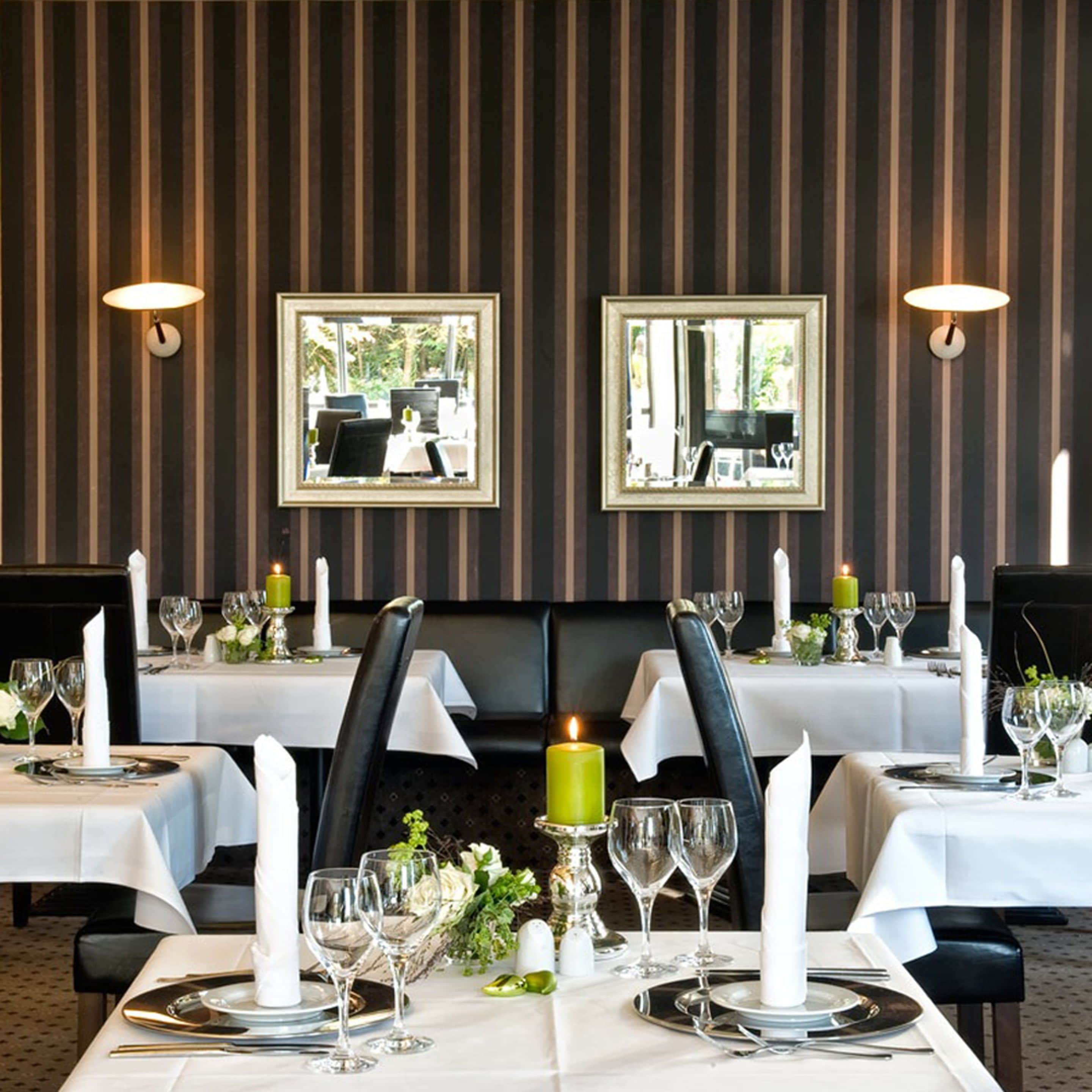 Un restaurant aux murs recouverts d’un papier peint à rayures, agrémenté de miroirs encadrés, et dont les tables sont dressées avec des nappes blanches, des bougies et des verres à vin.