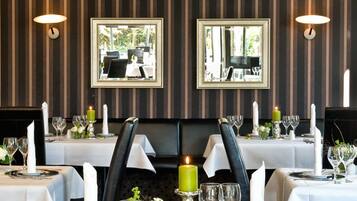 Un restaurant aux murs recouverts d’un papier peint à rayures, agrémenté de miroirs encadrés, et dont les tables sont dressées avec des nappes blanches, des bougies et des verres à vin.