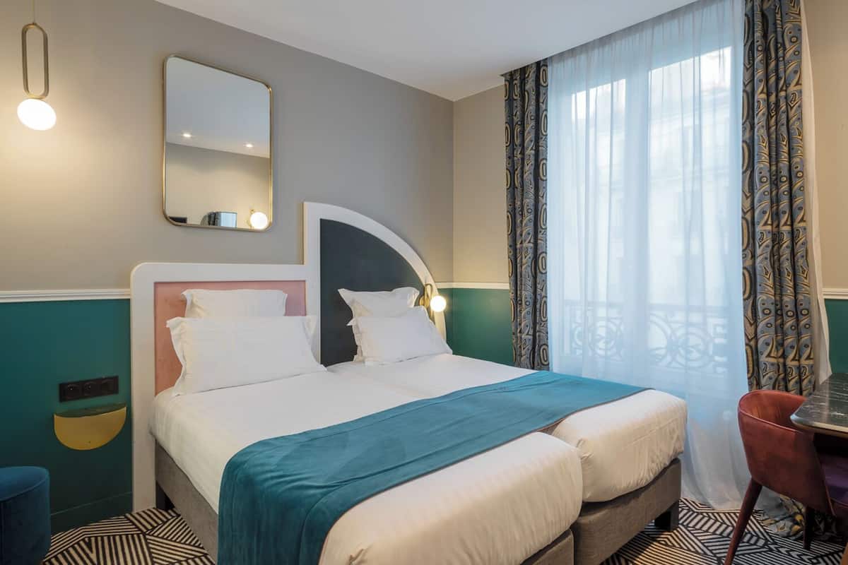 A hotel room with a bed, a mirror, a bedside table, a chair, and a window with curtains.
