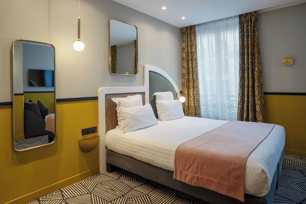 Una habitación de hotel con una cama grande, una mesita de noche, un espejo y una ventana con cortinas estampadas.