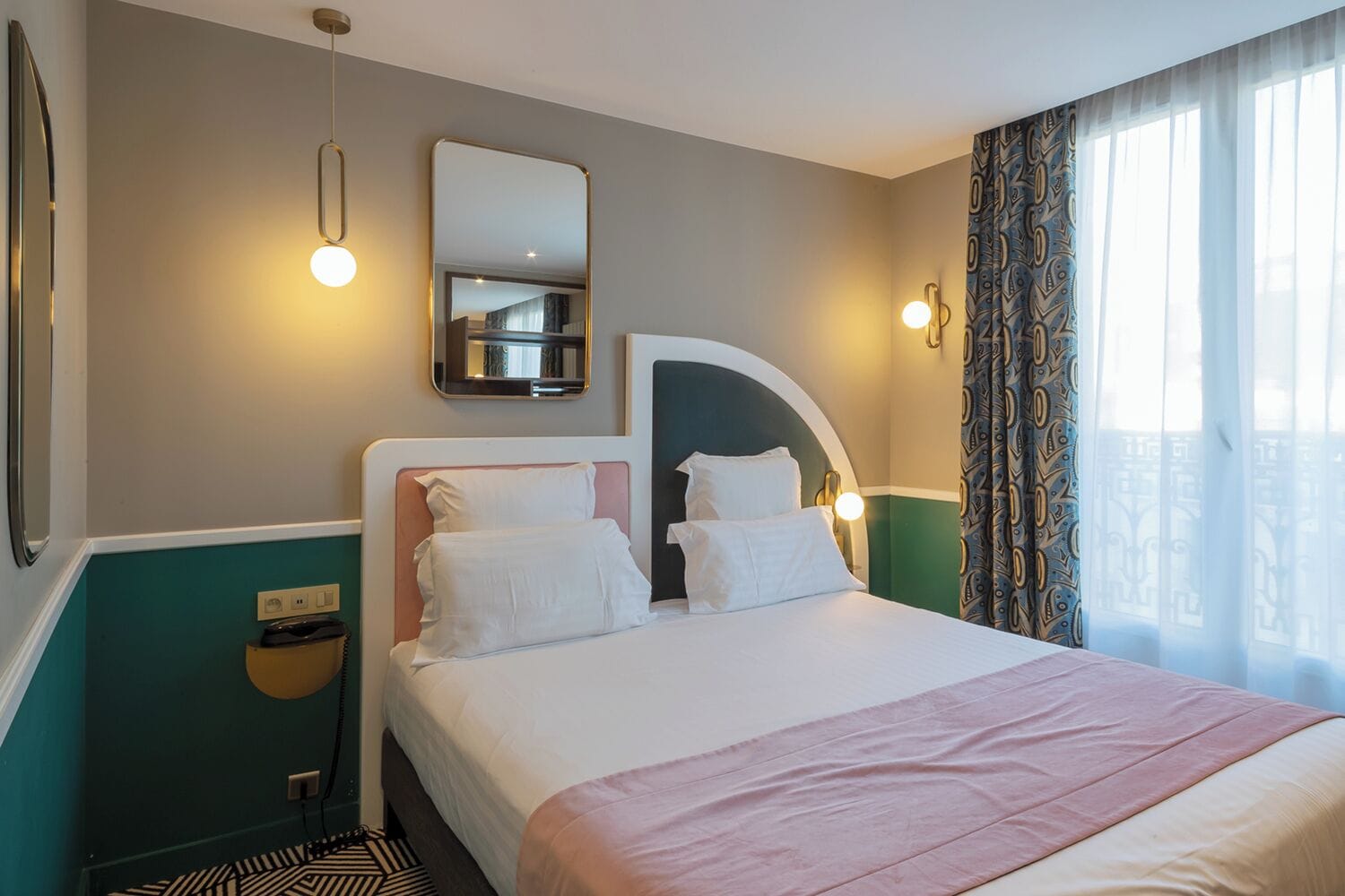 A hotel room with a bed, a mirror, a wall-mounted light, and a window with curtains.