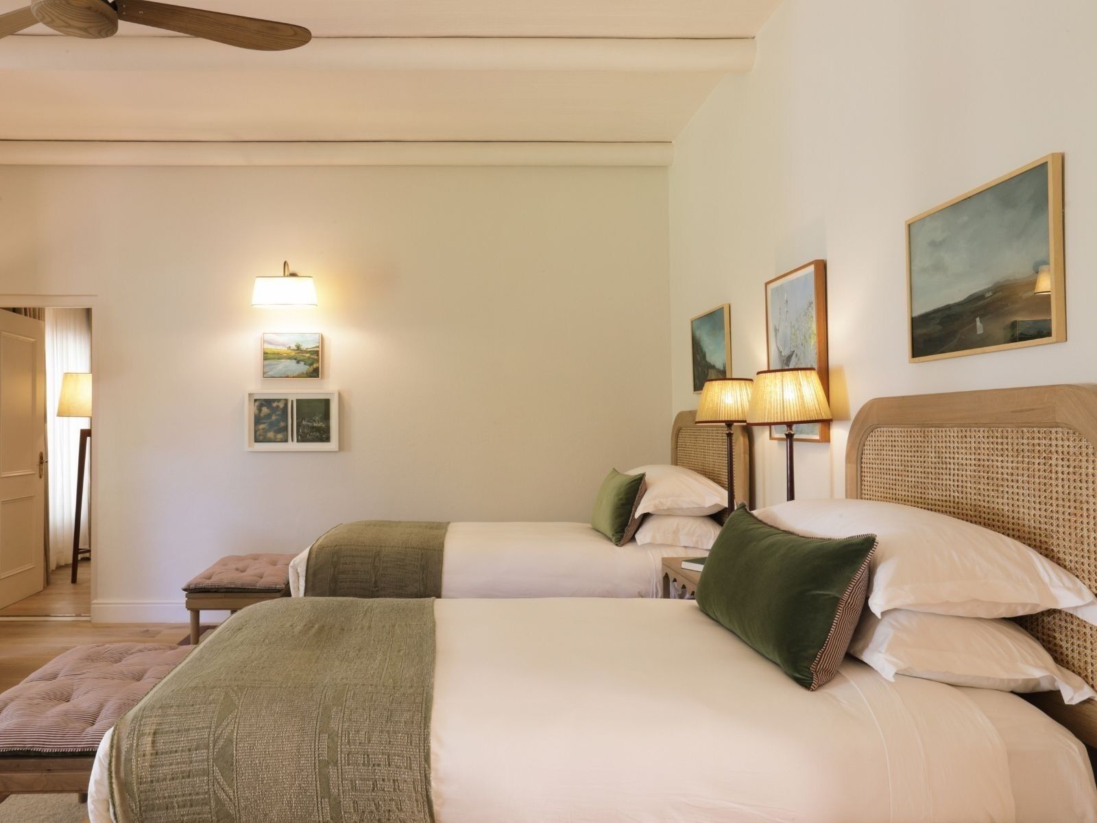 A hotel room with two beds, a ceiling fan, wall-mounted lamps, and framed pictures.