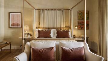 Een slaapkamer met een hemelbed, een nachtkastje met een lamp en een fauteuil met kussens.