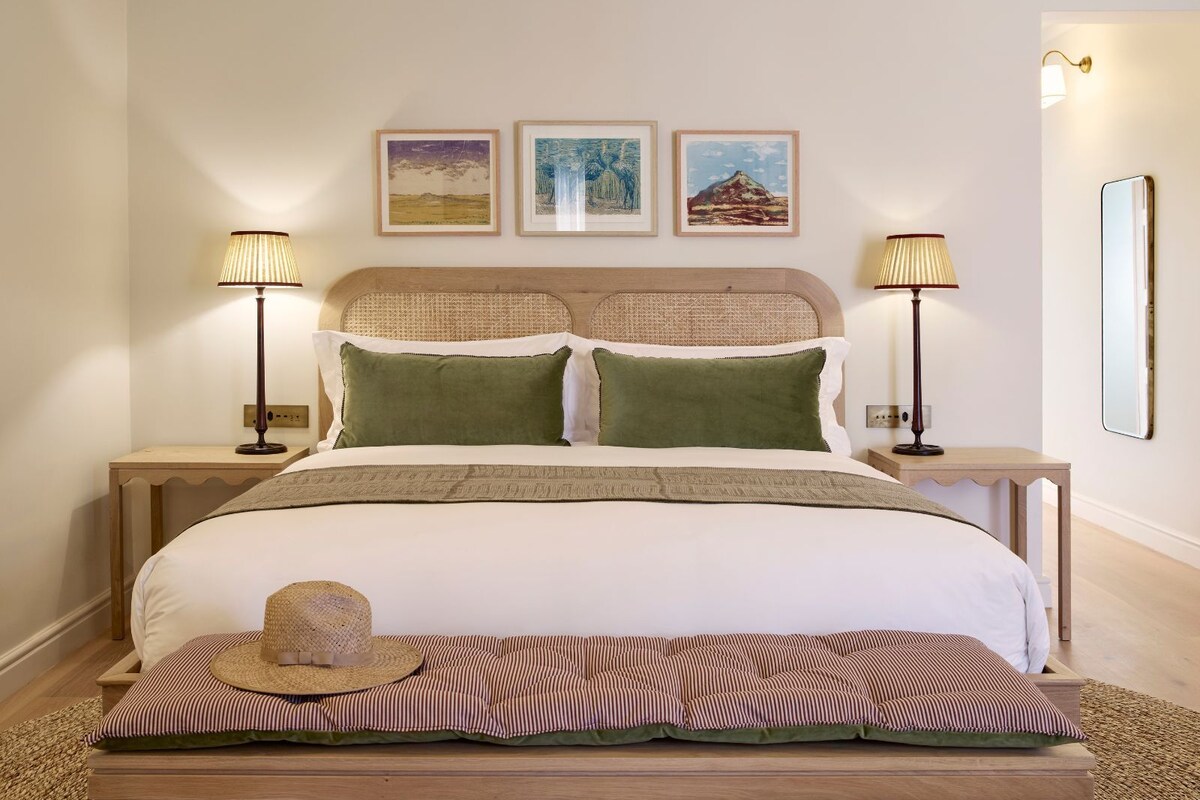 Una cama bien hecha con un cabecero, dos mesitas de noche con lámparas, un banco con un sombrero y tres cuadros enmarcados en la pared.