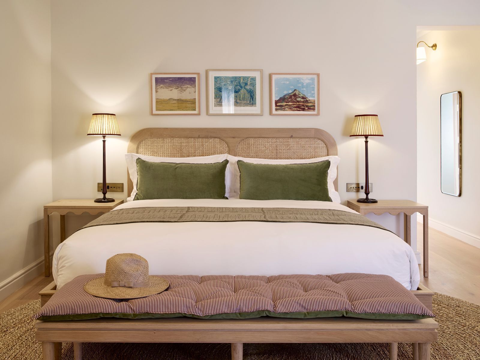 A neatly made bed with a headboard, two bedside tables with lamps, a bench with a hat, and three framed pictures on the wall.