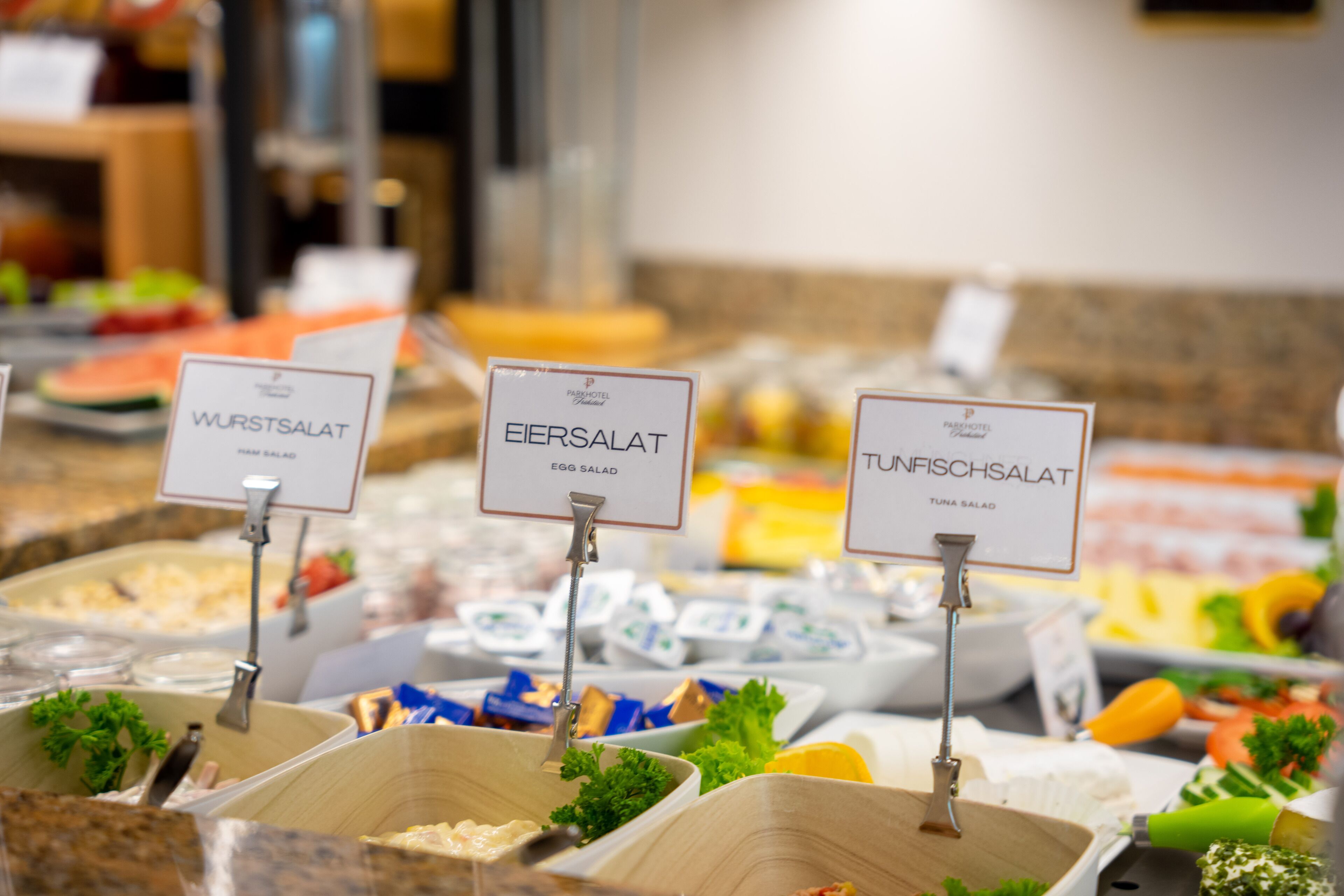 Ein Buffet mit Salatoptionen, darunter Wurstsalat, Eiersalat und Thunfischsalat, alles beschriftet.