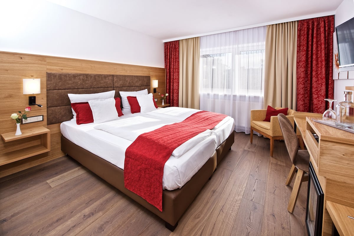 A hotel room with a large bed, a desk, and a chair. There is a TV on the wall and a window with curtains.