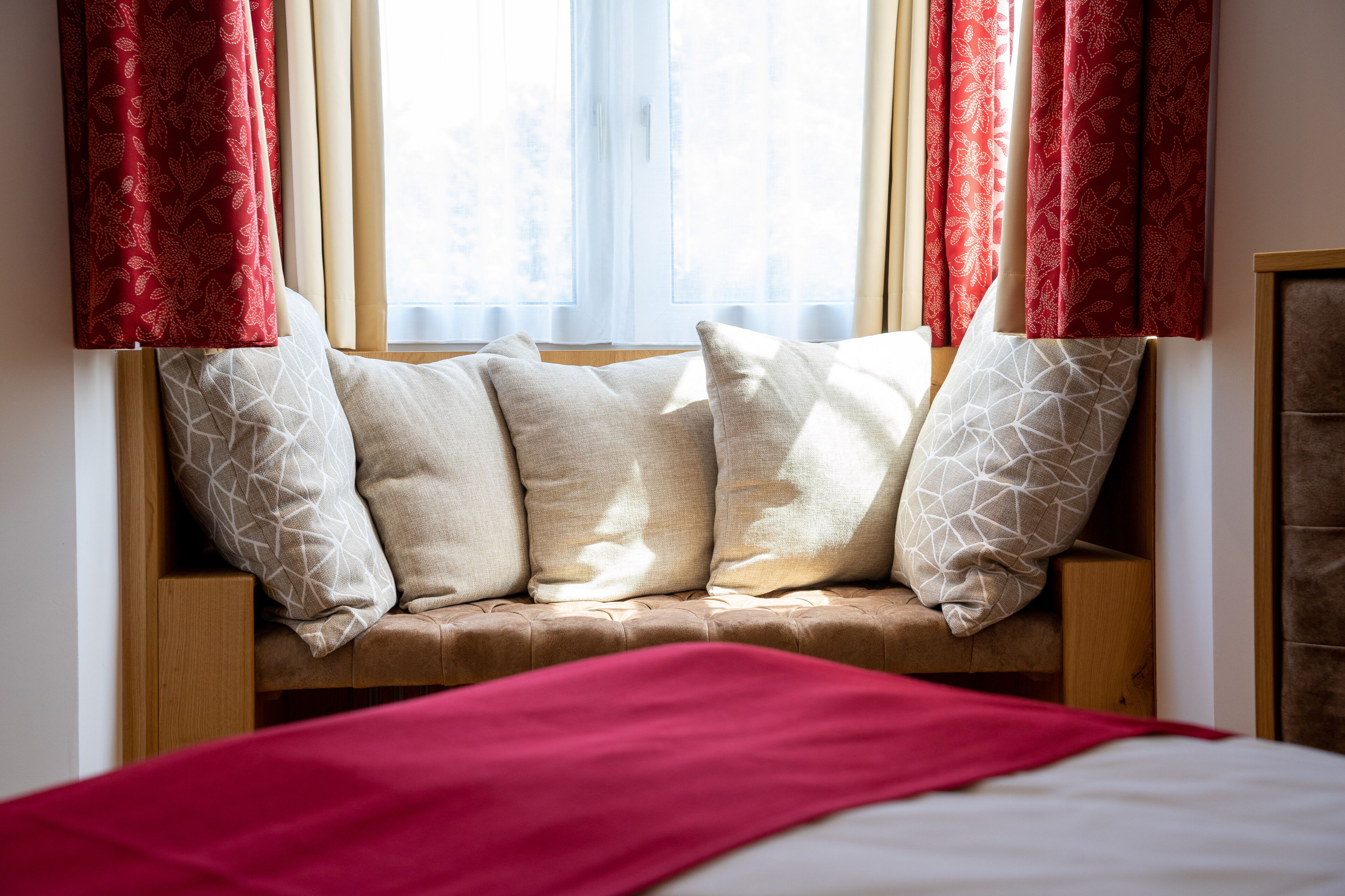 Quarto com assento à janela, almofadas bege e manta vermelha.
