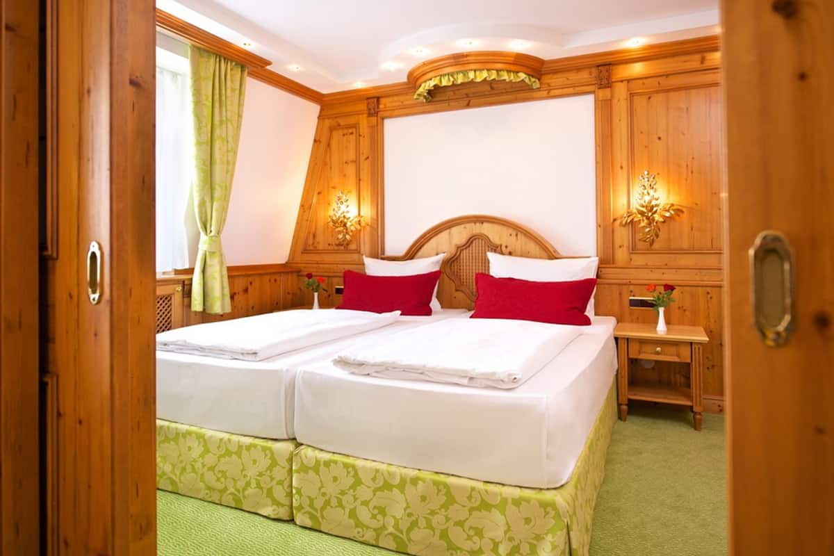 A double bed with white and green bedding, two red pillows, and a wooden headboard. There are two wall-mounted lamps and a small wooden nightstand with a vase of flowers.