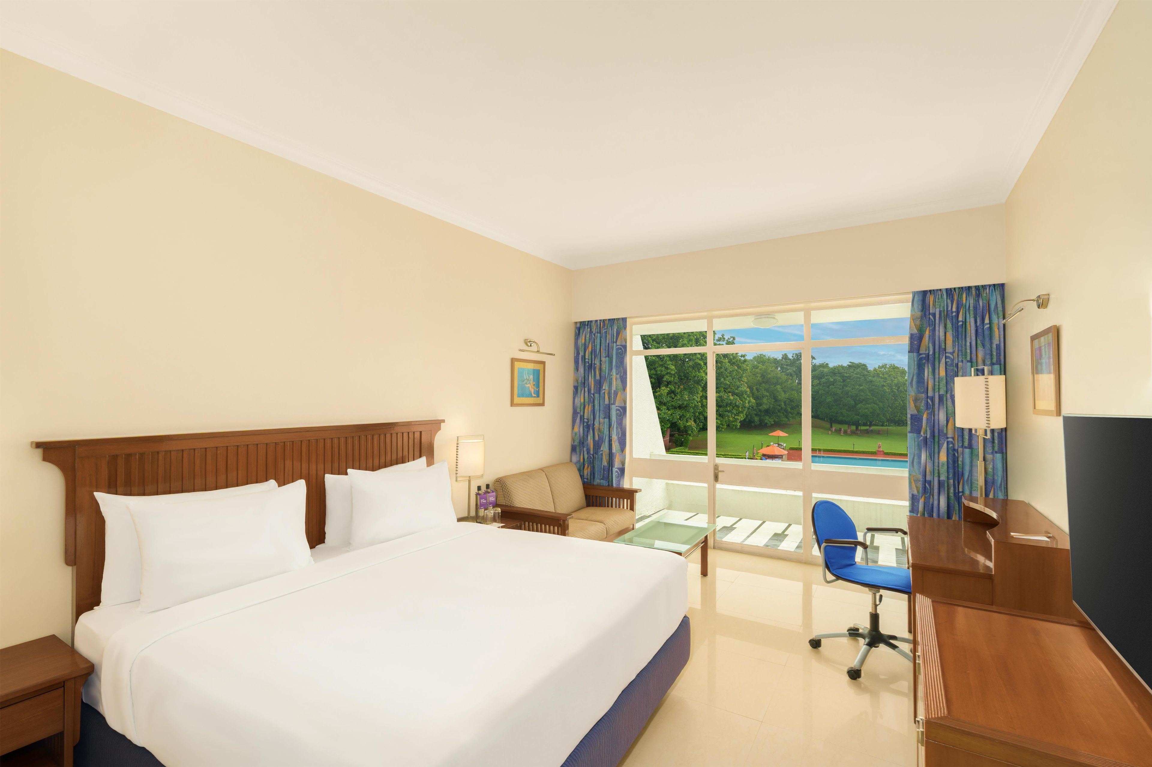 A hotel room with a large bed, a desk, a chair, and a view of a swimming pool and greenery.