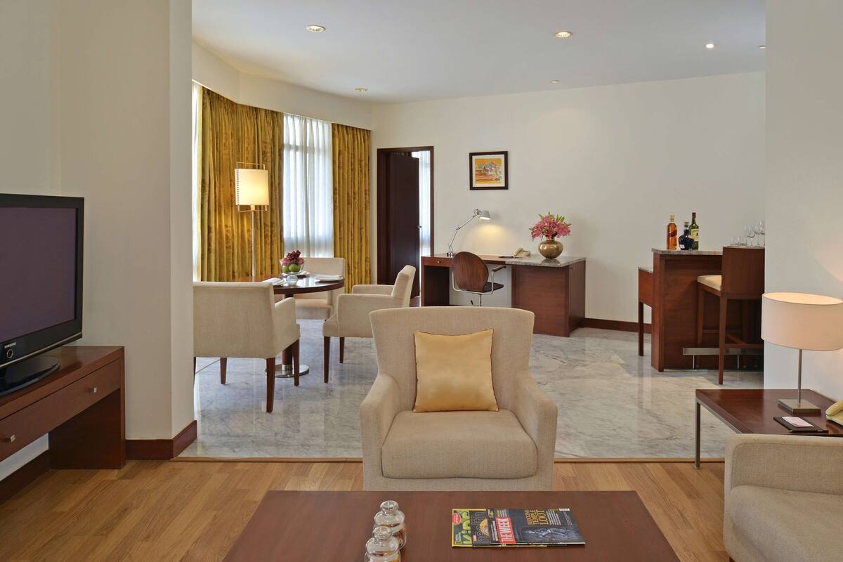 A hotel room with a TV, a dining area with a table and chairs, a desk with a lamp, and a sofa.