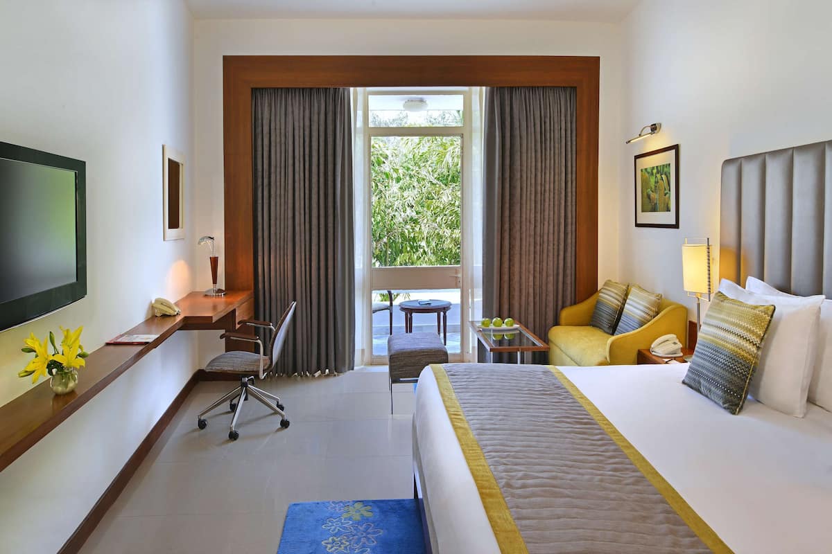 A hotel room with a bed, a desk with a chair, a TV, a window with curtains, and a view of greenery outside.