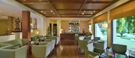 A spacious hotel lobby with a bar area, green upholstered seating, and a view of the garden.