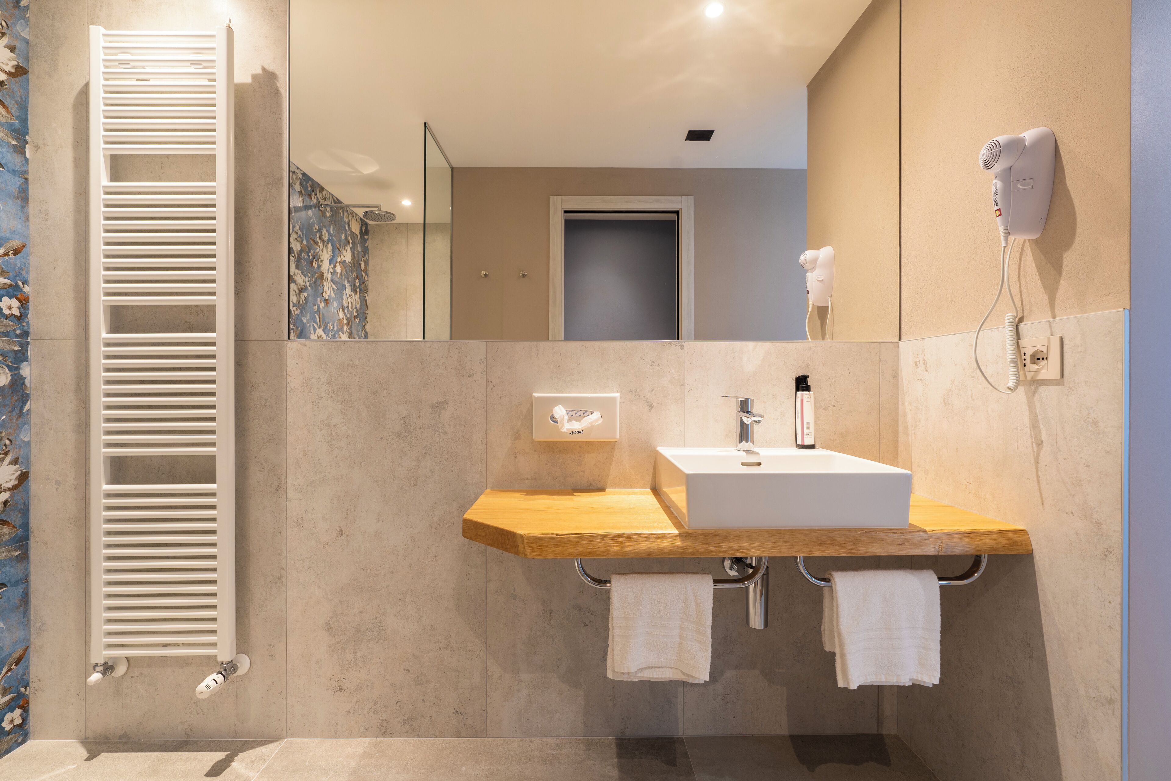 A modern bathroom with a wooden vanity, a white sink, a towel rack, a hairdryer, and a shower with a glass door.