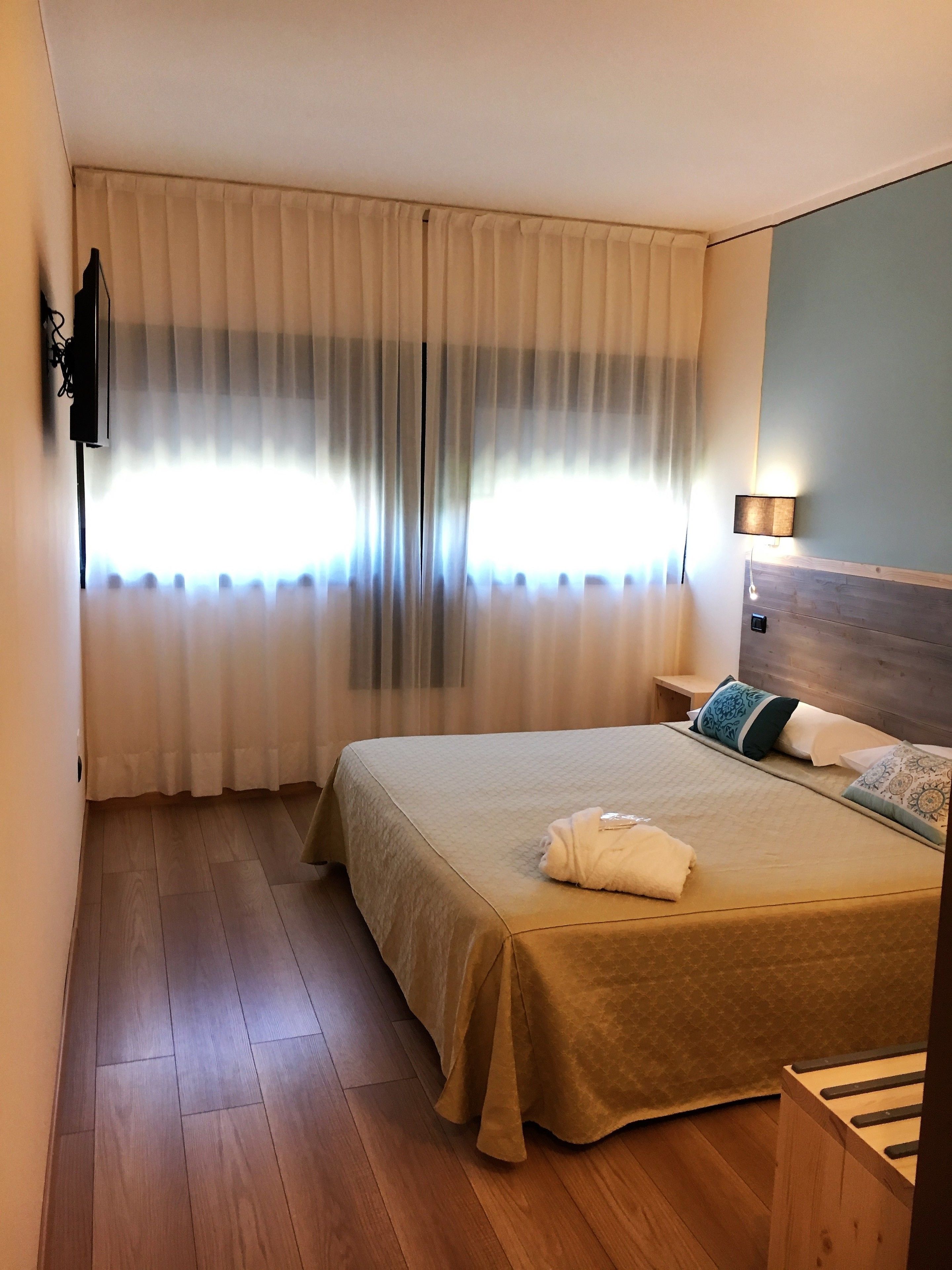 A hotel room with a bed, a wooden floor, a window with curtains, a wall-mounted TV, and a bedside lamp.