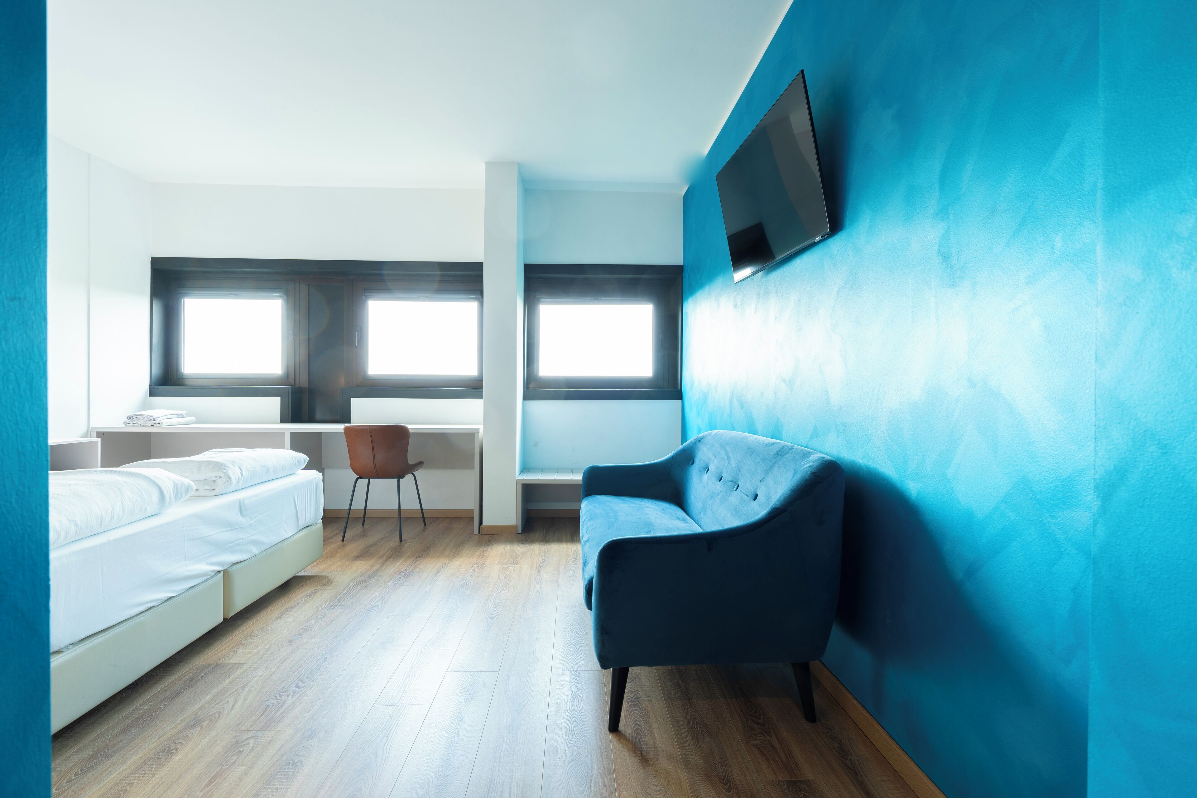 A modern hotel room with a bed, a desk, a chair, and a TV mounted on the wall.