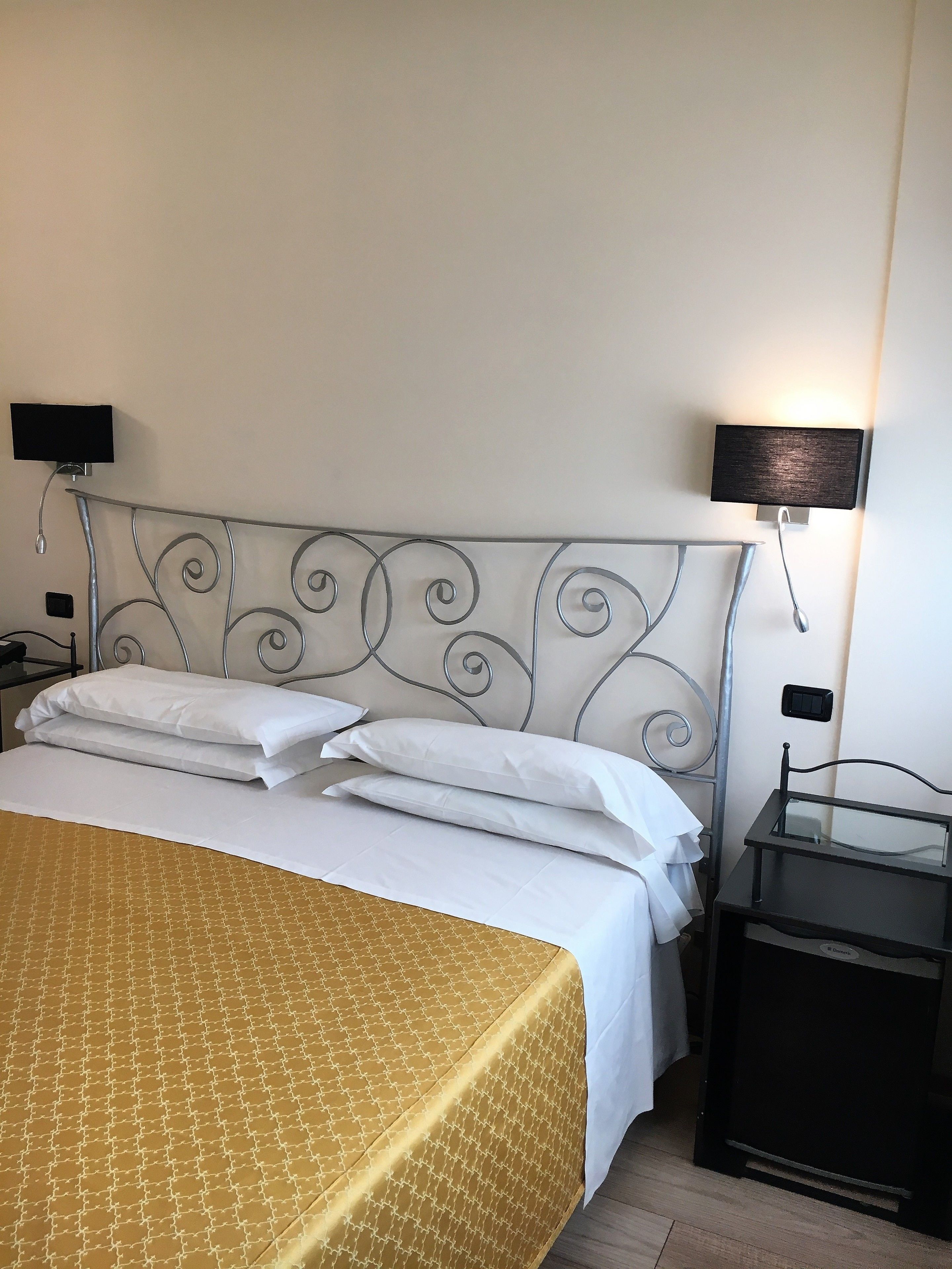A neatly made bed with white linens and a patterned bedspread, a metal headboard with decorative swirls, two bedside tables with lamps, and a small black nightstand.